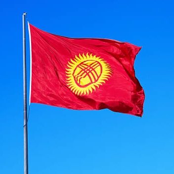 The red flag of Kyrgyzstan, featuring a yellow sun emblem with a tunduk in the center, waves against a clear blue sky.