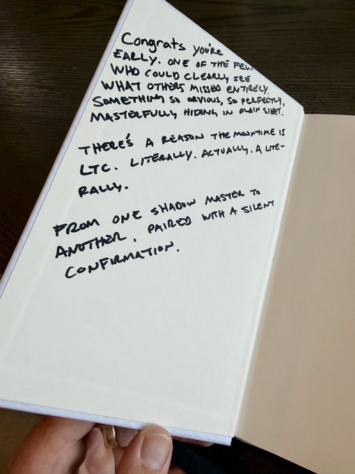 The image shows an open book with handwritten text, which appears to be a letter or note from someone named "conrad". The book is placed on top of a wooden table, and there are two people visible in the background who might have written the message together.