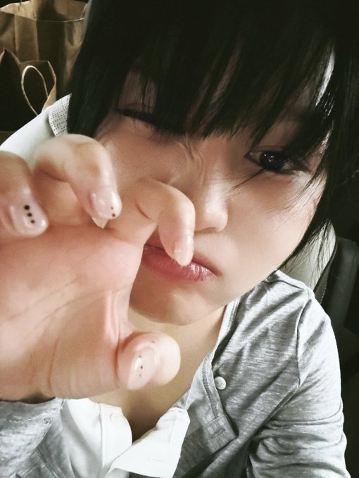A close-up selfie of a young woman with dark hair and bangs reaching her hand toward the camera, wearing a gray button-up shirt.