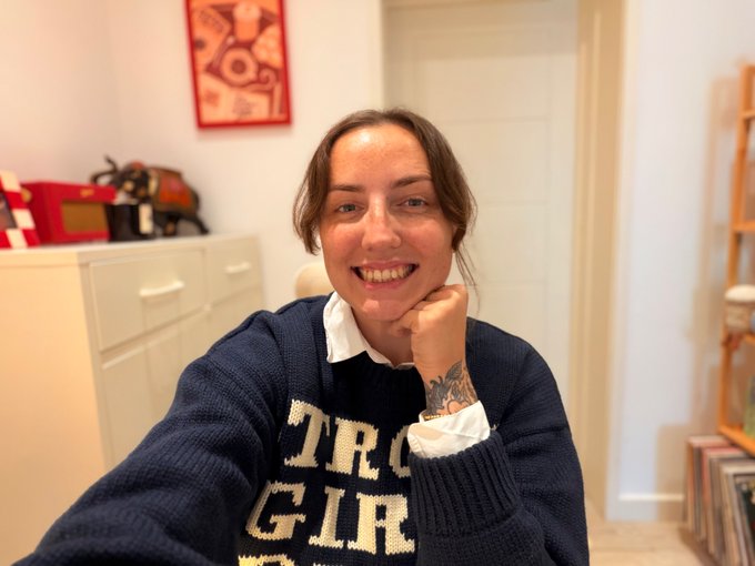 Selfie of a smiling young woman wearing a navy blue 'TROGIR' sweater over a white collared shirt, with a tattooed wrist, posing in a room with a red framed picture.