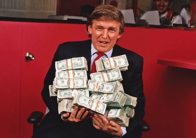The image appears to be AI-generated, depicting a Donald Trump lookalike holding several stacks of US dollar bills banded together, set against a red backdrop. It visually reinforces the post's theme of Trump's proposed $5,000 payout to Americans and the massive potential $1 trillion+ in liquidity that could flow into stocks and crypto markets ahead of the midterms.