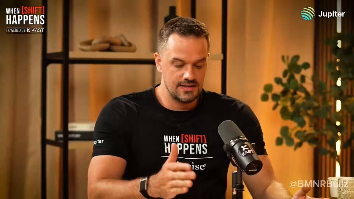The image features a man wearing a black t-shirt with white text that says "When SHIFTY HAPPENS Rise". He is sitting at a microphone, likely participating in an interview or podcast discussion about cryptocurrency. The background of the image shows a bookshelf filled with various items such as books and a potted plant.