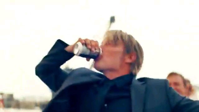 A blond man in a black jacket tilts his head back while drinking from a can outdoors.