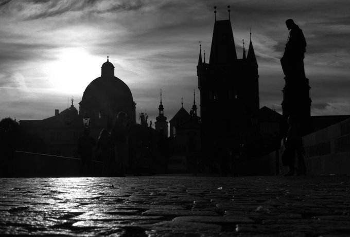 A black and white silhouette photograph shows a city skyline featuring Gothic towers and a domed building against a bright sunset, with people walking on a wet cobblestone bridge in the foreground.