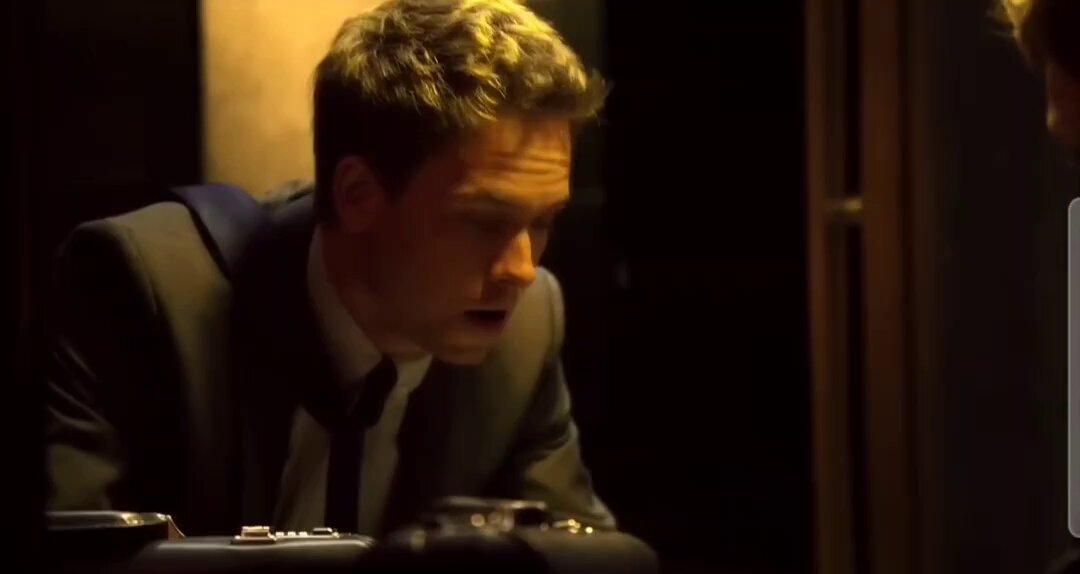 The image shows a man sitting at a desk with his head down, looking at something on the table or possibly a camera. The background appears dark, which contrasts with the bright light shining on the man's face. There are two other people visible in the scene, one of them wearing a tie and another person standing behind him. The image also contains text that reads "focus on what is in the image and how it relates to the post".