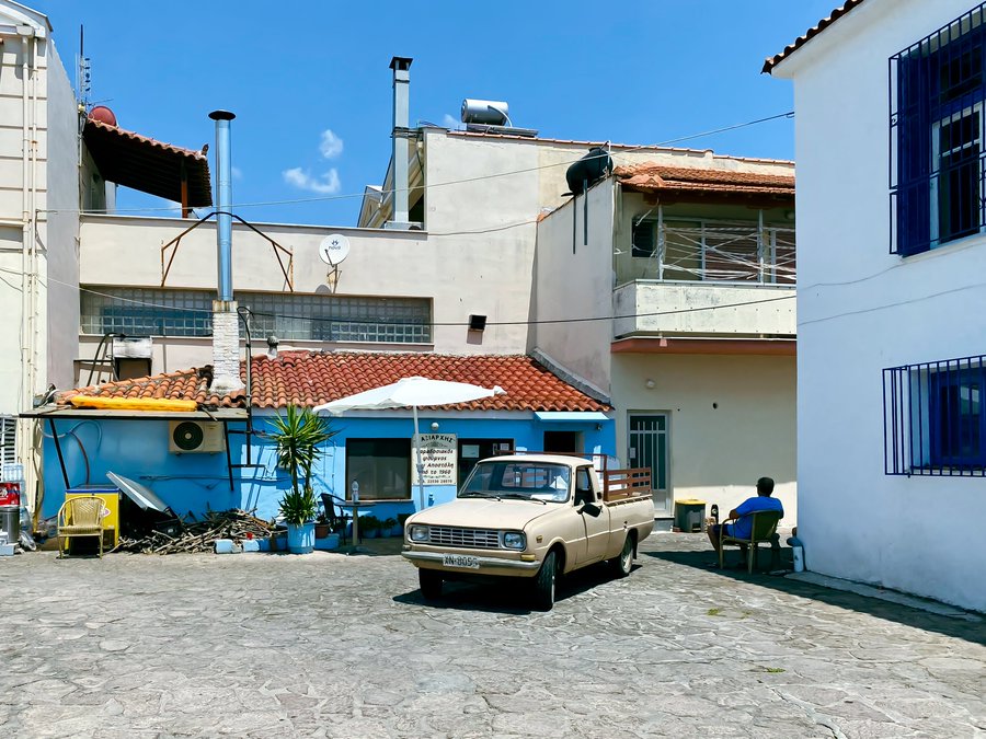 A sunlit Mediterranean-style village courtyard with a cobblestone ground, featuring a vintage white pickup truck parked near a blue building with a 'ΔΙΑΦΗΜΙΣΗ' (advertising) sign, while a man relaxes in a plastic chair nearby.