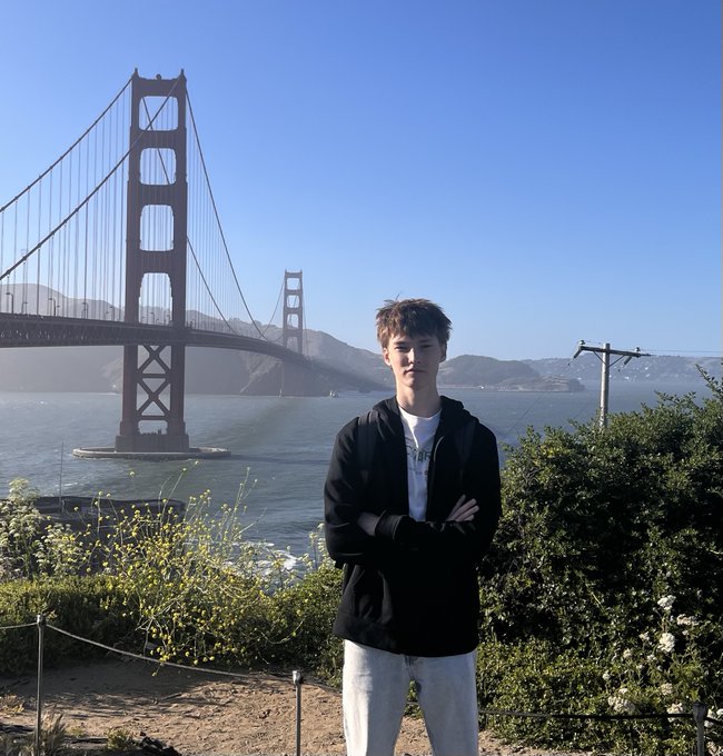 In the image, there is a young man wearing black clothing who stands in front of a bridge. He has his arms crossed and appears to be posing for a photo or selfie. The backdrop includes the iconic Golden Gate Bridge over water below him. The caption provided seems unrelated and could potentially confuse viewers about what this photograph actually depicts.