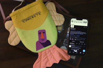 A felt craft project featuring a purple character with the text "CONCRETE" sits on a pink towel next to a smartphone displaying a QR code and social media profile.
