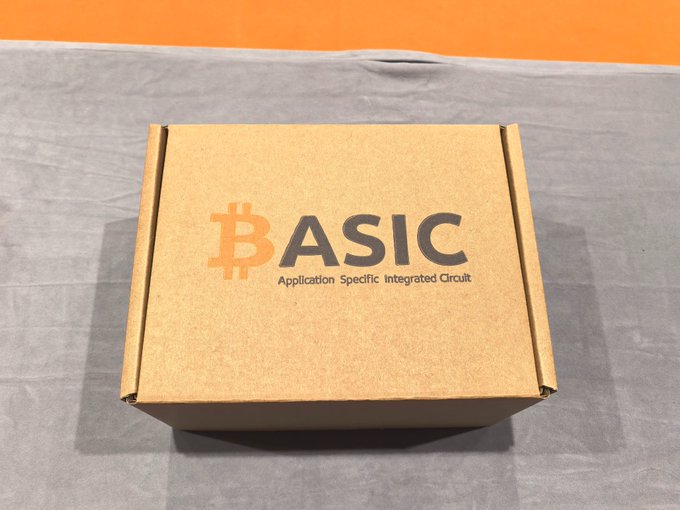 A cardboard shipping box printed with 'BASIC - Application Specific Integrated Circuit' alongside a Bitcoin logo, sitting on a gray surface against an orange background.