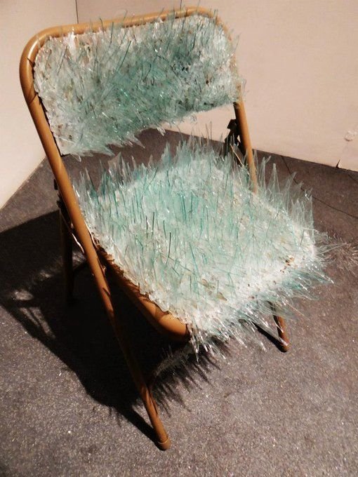 A wooden folding chair covered in numerous sharp, light blue glass shards sticking upward from the seat and backrest.