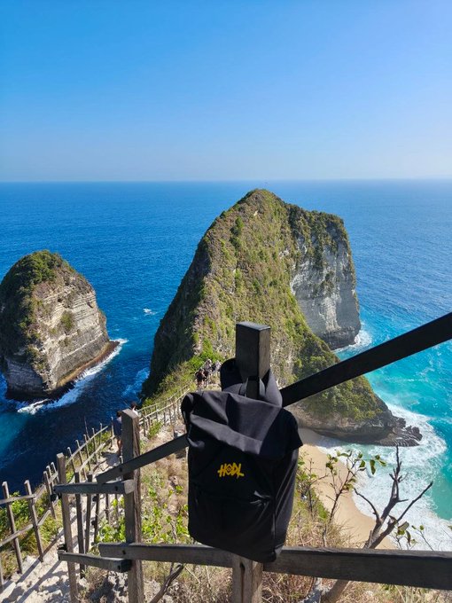 A black tote bag with "#HODL" printed in yellow hangs on a wooden railing overlooking the iconic Kelingking Beach cliffs of Nusa Penida, blending crypto culture with a breathtaking ocean view for a "GM #HODL" post.