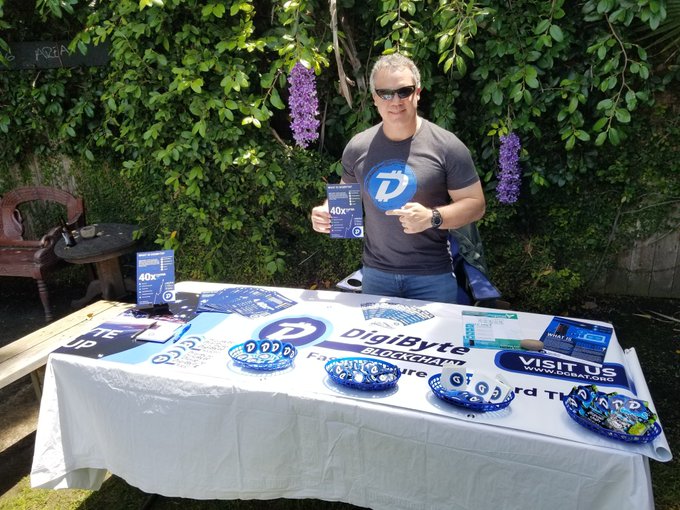 A man wearing sunglasses and a Digibyte-branded t-shirt stands behind a table displaying DigiByte promotional merchandise, stickers, and 'VISIT US' materials at an outdoor event with wisteria vines in the background.