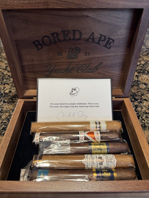 An open Bored Ape Yacht Club 2021 wooden cigar box containing several cigars and a card reading 'Five years deserves a proper celebration. This is ours. Five years. Five Cigars. One box. Bored Ape Yacht Club.' signed by Yuga Labs.