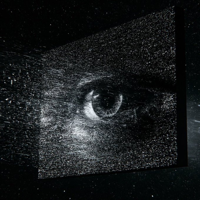 A black and white image resembling a screen or projection displays a bright, particle-like burst pattern that forms the shape of a human eye in the center.