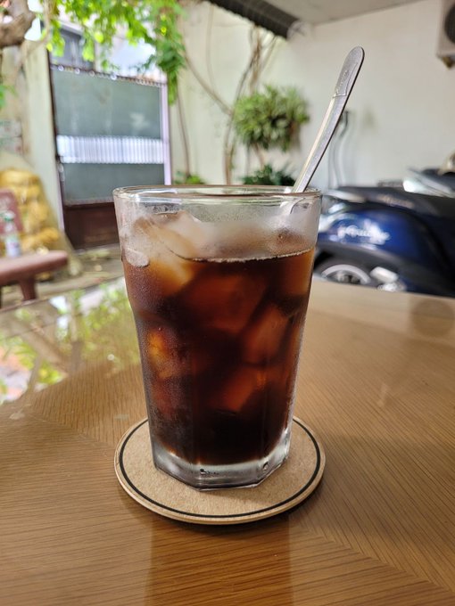 A glass of iced coffee with a spoon sits on a coaster atop a wooden table at an outdoor cafe, with greenery and a parked motorcycle visible in the background.