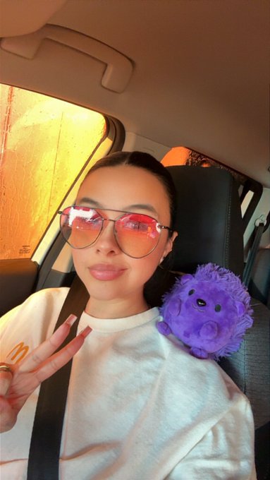 A selfie of a woman wearing reflective pink-tinted sunglasses and a white McDonald's t-shirt, with a small purple plush toy in the car.