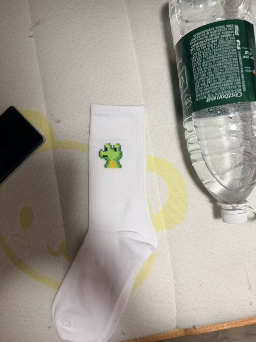 A pair of white socks featuring a pixelated green cactus character lies on a bed next to a Gerolsteiner water bottle and a smartphone, matching the post's mention of "white socks from GMGN."