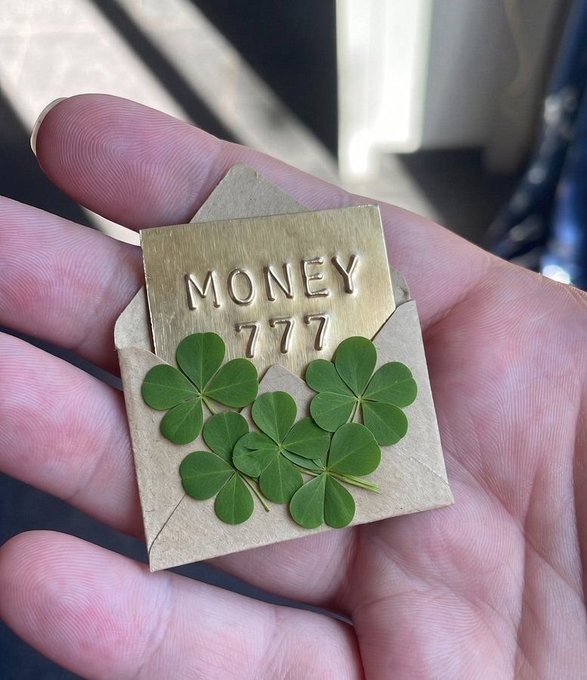 A hand holds a small brass envelope charm engraved with 'MONEY 777', filled with green shamrock clovers.