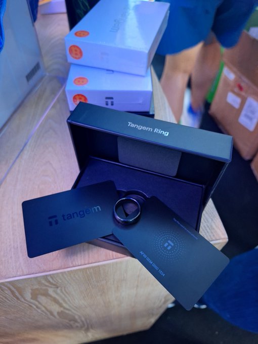The Tangem Ring, a sleek black crypto hardware wallet, is displayed in its open packaging alongside the "tangem" branded cards and ring holder. Stacked boxes with orange seals sit in the background, ready for shipping or unboxing.