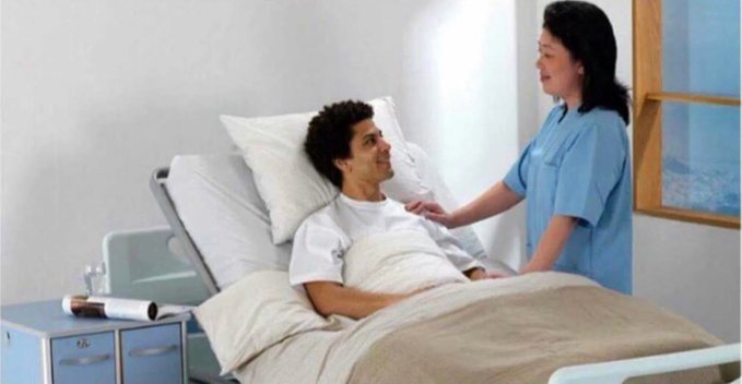 The image shows a woman standing next to a hospital bed with a male patient lying down on it. The woman appears to be talking or comforting the patient while they are both facing each other. A window can also be seen in the background, suggesting that this scene takes place within an indoor setting like a hospital room.