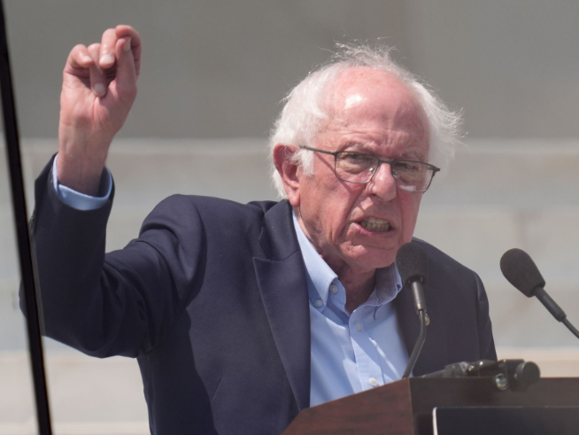Bernie Sanders speaking passionately at a podium with two microphones, wearing a navy blazer and light blue shirt, raising his fist.