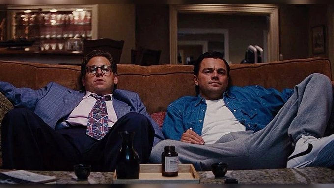The image is a famous scene from *The Wolf of Wall Street* showing Jonah Hill and Leonardo DiCaprio slouched on a couch with blank expressions, perfectly capturing the post's "Gm Let's make a Bag" vibe of detached, laid-back confidence.

Caption: Two men in business-casual and casual attire recline on a couch with deadpan expressions, exuding a relaxed, almost sedated aura beside a small bottle on the coffee table.