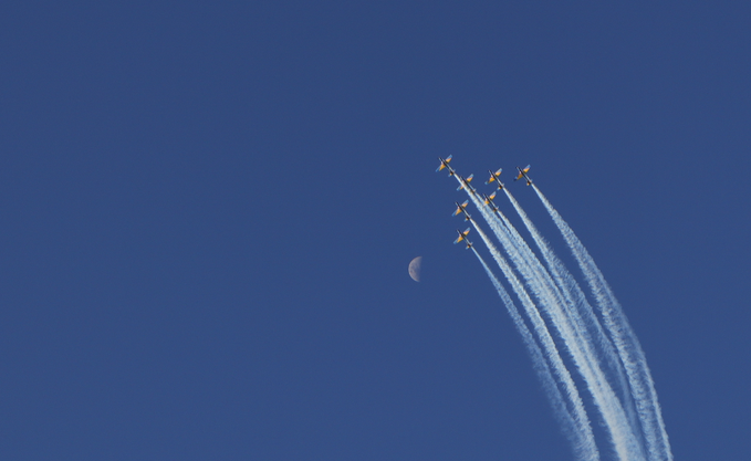 Six jets flying in formation leaving white contrails against a deep blue sky, with a crescent moon visible in the background.