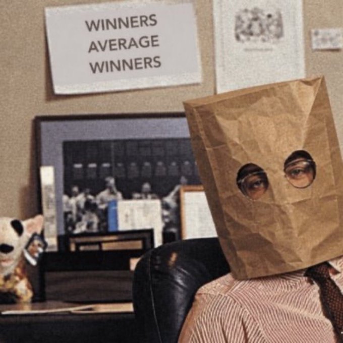 A man sits in an office chair wearing a brown paper bag over his head with eye holes cut out, beneath a motivational poster reading "WINNERS AVERAGE WINNERS," while a cat figurine and framed sports photos sit on a shelf nearby.
