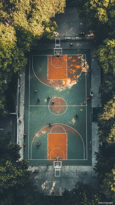 An aerial view of a green and orange outdoor basketball court surrounded by trees, with several people playing and walking around, watermarked 'pixelstalk.net'.
