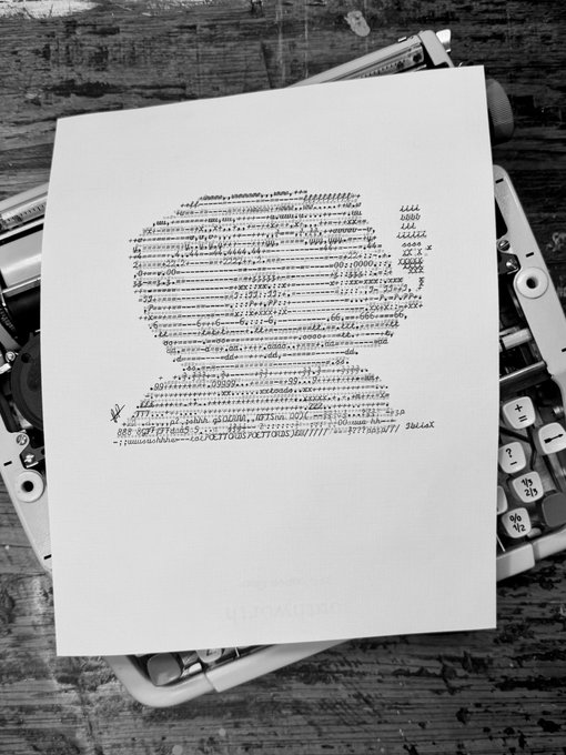 A black and white photograph of a printed paper containing what appears to be ASCII art or computer code, resting on a wooden surface with a keyboard.