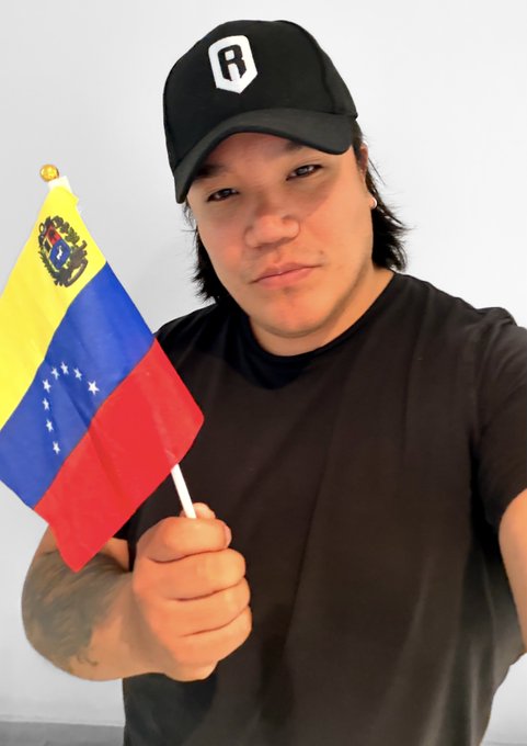 A man wearing a black "R" cap and black t-shirt holds a small Venezuelan flag, dedicating his Axie Selfie to his Venezuelan friends in support of the Ronin Network community's fundraising effort for @Tita_KIND.