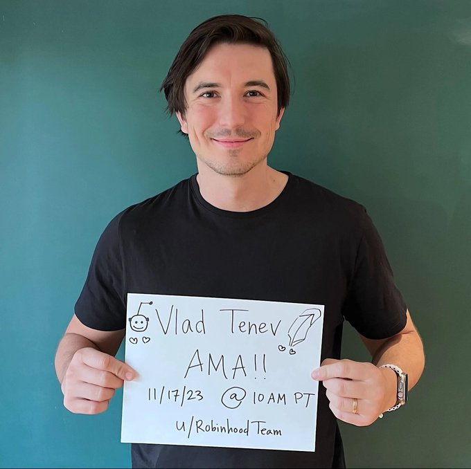 A man holds a handwritten sign reading "Vlad Tenev AMA!! 11/17/23 @ 10AM PT u/Robinhood Team," promoting an Ask Me Anything session with the Robinhood CEO tied to the discussion about launching Robinhood Chain onchain.