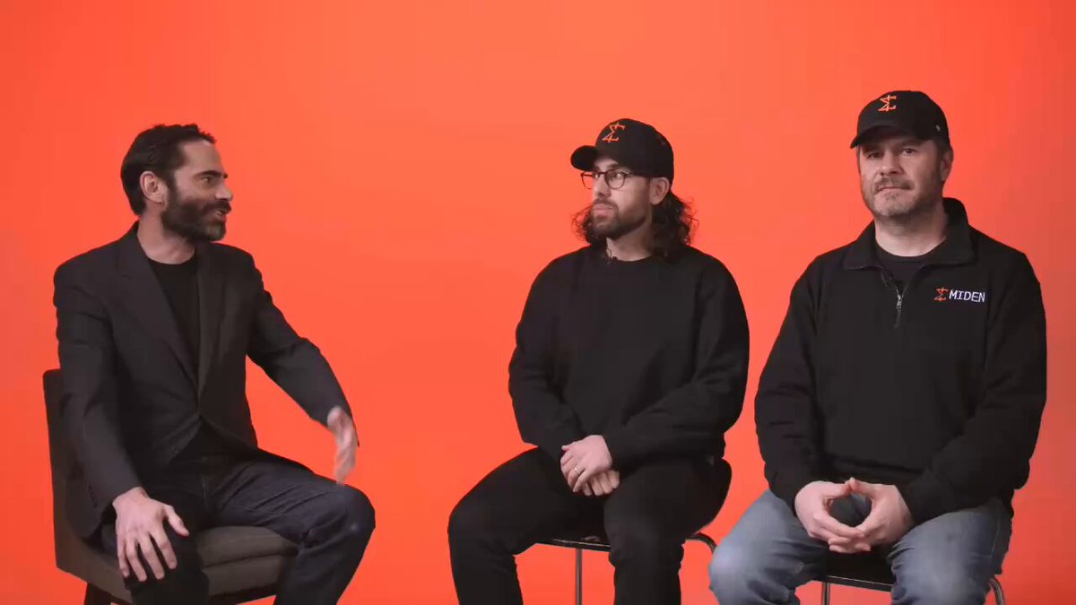 The image features three men sitting next to each other, with one man wearing a black suit jacket and another two men wearing baseball caps. The background of the image is orange, which contrasts with the gray wall behind them. There are no texts or additional objects in the image, and the focus remains on the people seated together.