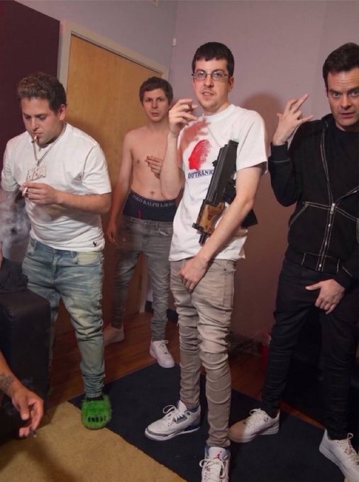 A group of four young men stand in a room; one on the left wears a white t-shirt and a large chain, another in the center wears a white t-shirt with a red logo and holds what appears to be a firearm, a shirtless man stands in the background, and a fourth man on the right wears a black jacket and gestures with his hand.