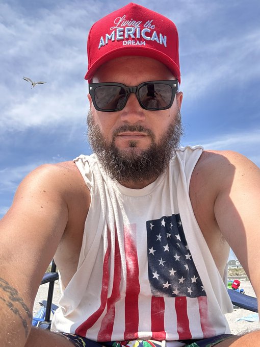 A bearded man in sunglasses wears a red 'Living the AMERICAN DREAM' cap and an American flag tank top while sitting outdoors at the beach with a seagull in the background.