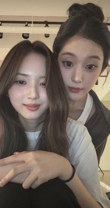 Two young women pose closely together for a selfie, with the woman on the left having her arms wrapped around the other in an affectionate embrace, both looking directly at the camera with soft, neutral expressions.