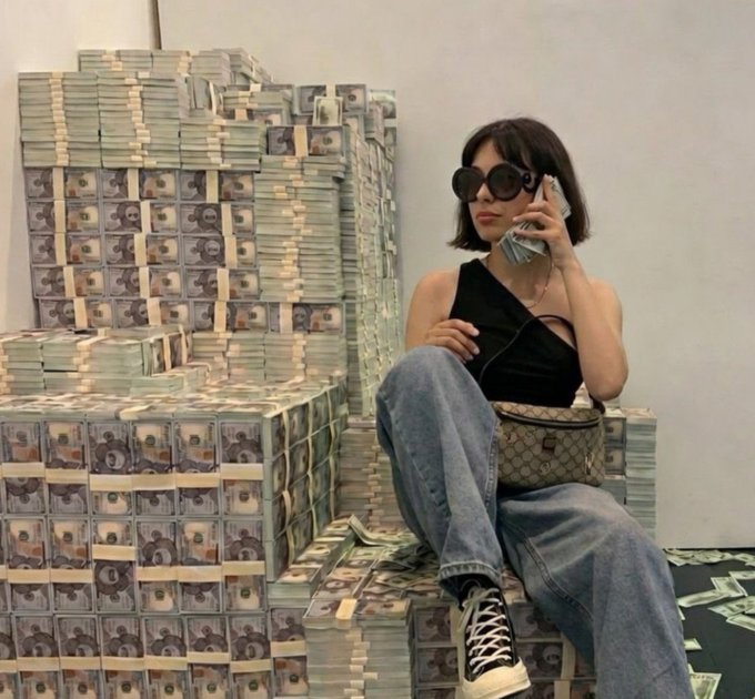 A woman wearing dark round sunglasses and a black top poses with a phone on her ear and a Gucci bag, sitting beside massive stacks of banded US dollar bills.