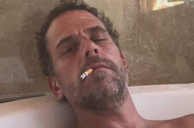 A close-up of a man with a graying beard relaxing in a bathtub with a lit cigarette in his mouth.