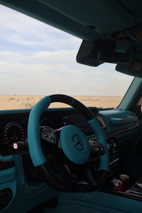 A turquoise Mercedes-Benz interior with a Brabus steering wheel overlooks a desert landscape. The luxury customization aligns with the "GM (get money)" theme, symbolizing wealth, success, and high-end automotive prestige.
