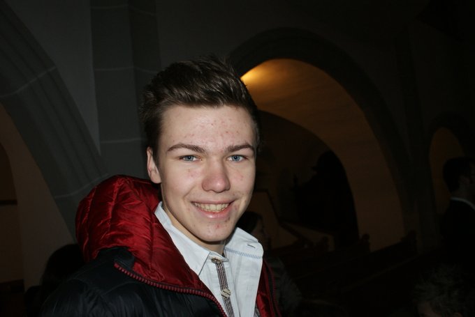 A close-up portrait of a smiling young man with brown hair wearing a red and black jacket over a white shirt, standing in what appears to be a dimly lit church or arched interior.