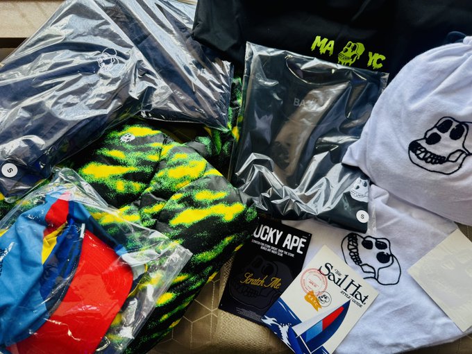 A collection of Bored Ape Yacht Club merchandise is spread out, including a white cap featuring the iconic ape skull logo and various items in plastic packaging, with "Bored Ape Yacht Club" branded tote bags, a "Mutant Ape" label, and a "Scratch Me" tag visible. The premium packaging and presentation of the items—including the free Mutant Tote and cap—reflects the top-notch unboxing experience mentioned in the post.