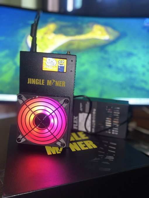 A 'JINGLE MINER' cryptocurrency mining device with a glowing pink and orange fan sits on top of its box, with a monitor displaying a map in the background.