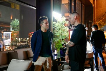Two men in dark clothing stand talking at a dimly lit restaurant or bar at night, with one in a blue blazer and the other in a black shirt.