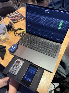 A laptop sits on a wooden table displaying a trading interface with green data rows, alongside a black calculator, a smartphone showing a financial app, and a small card reader, suggesting a workspace for financial tracking or trading.