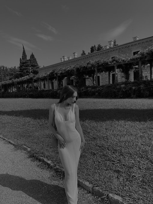 A black and white photo of a pregnant woman in a light slip dress walking along a path in front of a long, ivy-covered building.