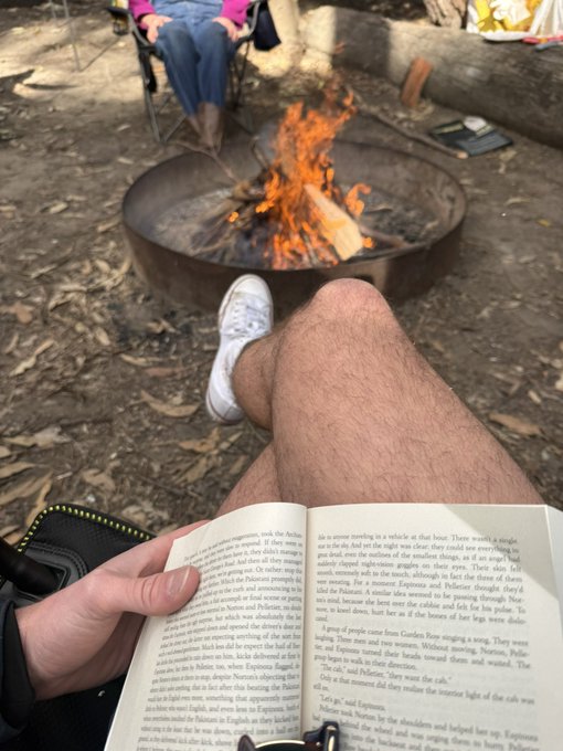 A person relaxes by a crackling campfire, reading a paperback book while wearing white sneakers, capturing the serene contrast between modern digital life and a peaceful outdoor escape. The image humorously underscores the caption's sentiment—highlighting how even in a tranquil setting like this, the temptation of trading perpetual futures ("perps") on a phone lingers, intruding on simple moments of unplugged leisure.