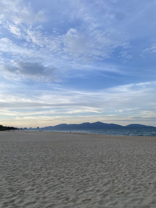 A serene beach scene with light-colored sand stretching toward a calm sea, with distant mountains on the horizon under a partly cloudy sky.