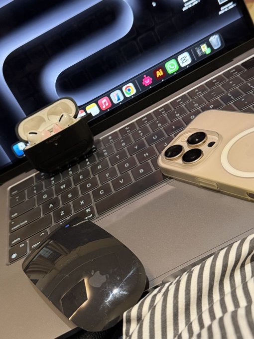 A desk setup features an open laptop with a 'MacBook Pro' keyboard, a pair of white wireless earbuds in their case, an iPhone in a tan case, and a black Apple Magic Mouse.