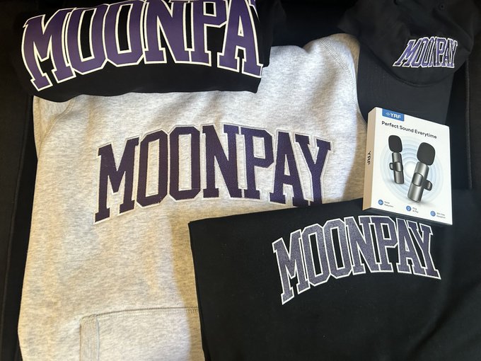 A collection of Moonpay-branded merchandise, including a grey hoodie and black items featuring purple 'MOONPAY' text, alongside a box for 'M2' wireless earbuds.