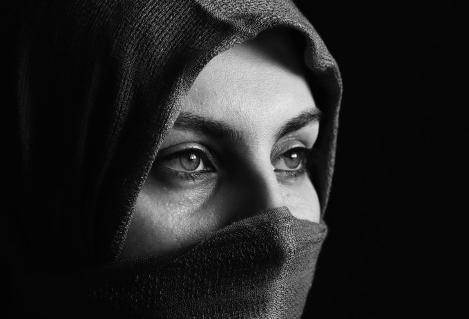 A black and white close-up portrait of a person wearing a dark hood and a face covering that reveals only their striking eyes.